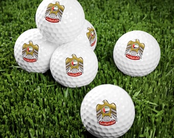 May include: Six white golf balls with the flag of the United Arab Emirates printed on them. The balls are arranged in a pyramid shape on a green surface.