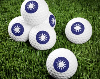 May include: Six white golf balls with a blue star and sun design on each ball. The balls are arranged in a pyramid shape on a green grassy surface.