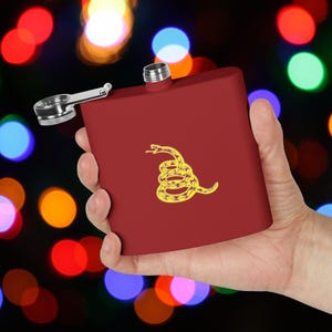 May include: A red metal flask with a yellow snake design. The snake is coiled and has its head raised. The flask has a silver cap and a hinged lid.