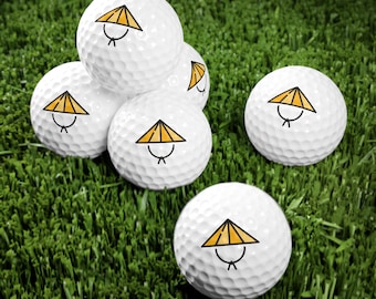 May include: White golf balls featuring a yellow and black straw hat design. Several balls are arranged on a green surface, some stacked and others scattered. The golf balls have a dimpled surface.