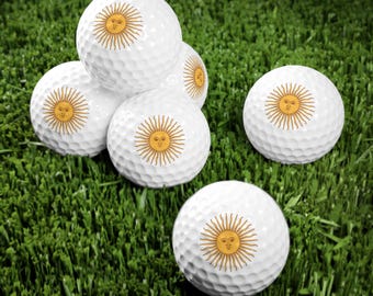 Pode incluir: Seis bolas de golfe brancas com um desenho de sol amarelo em cada bola. O sol tem um rosto e é rodeado por raios. As bolas de golfe estão dispostas em forma de pirâmide sobre uma superfície verde coberta de grama.