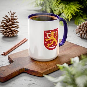 May include: White ceramic mug with a blue rim and handle. The mug features a graphic of the Finnish coat of arms, a red shield with a golden lion holding a sword.