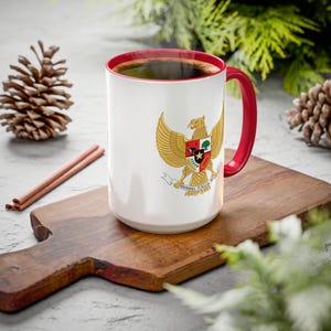May include: White ceramic coffee mug with a red rim and handle. The mug features a colorful image of the Indonesian coat of arms, a golden eagle with a shield and the text "Bhinneka Tunggal Ika".
