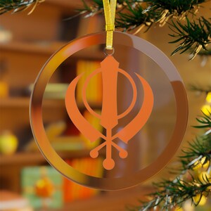 Puede incluir: Un adorno redondo de vidrio con una cinta dorada y un símbolo Khanda naranja, un símbolo religioso del sijismo.