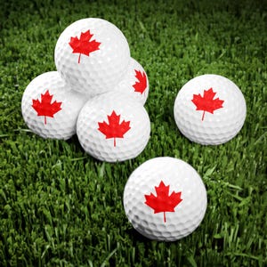 May include: Five white golf balls with a red Canadian flag design on each ball. The golf balls are on a green artificial grass surface.