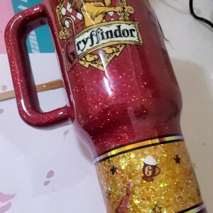 May include: A red glitter tumbler with a handle, featuring the Gryffindor crest and the word "Gryffindor". The bottom section has a gold glitter band with stars and a letter "G".