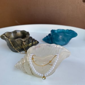 Puede incluir: Tres cuencos decorativos con forma de concha en varios colores: bronce, turquesa y blanquecino. La concha blanquecina presenta un collar dorado con una hilera de perlas y un pequeño colgante de gema transparente. Los cuencos están dispuestos sobre una superficie blanca.