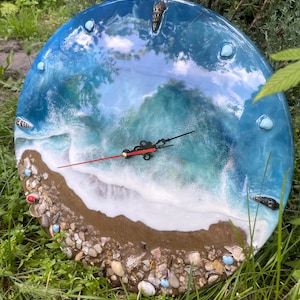 Könnte beinhalten: Runde Wanduhr mit Strandmotiv. Das Zifferblatt zeigt eine blau-weiße Ozeanszene mit Sandstrand, Muscheln und Kieselsteinen. Die Zeiger der Uhr sind schwarz mit einem roten Sekundenzeiger.