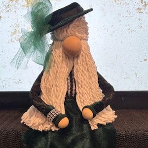 May include: A handmade gnome doll with a green dress, black sleeves, and a black hat. The gnome has a long white beard, wooden nose, and green boots. A green bow is attached to the hat. The doll is sitting on a dark surface.