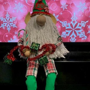 May include: A Christmas gnome wearing a green hat with a red and white candy cane, holding red and green gifts and a candy cane. The gnome is wearing plaid pants and green socks with white pom-poms.