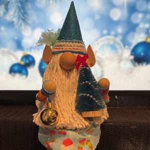 May include: A Christmas gnome doll with a teal glitter hat, wooden nose, and a white beard. The gnome holds a small Christmas tree and a gold ornament. The gnome wears a blue patterned outfit with red and green accents.