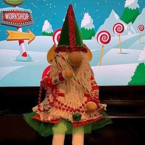 May include: A Christmas gnome doll wearing a green and red hat, a white sweater with red beads, a green skirt, and white boots with red and green accents. The gnome is holding a candy cane.