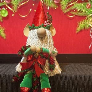 May include: A Christmas gnome doll with a red cone hat, white beard, and green and red outfit. The gnome is holding a green wreath with red bows and is wearing red and white patterned boots. Festive holiday decor.