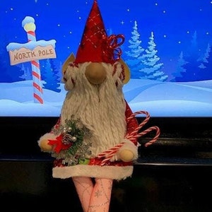 May include: A red and white gnome with a white beard and a red hat. The gnome is holding a candy cane and a small Christmas tree. The gnome is sitting on a black surface with a blue and white background.