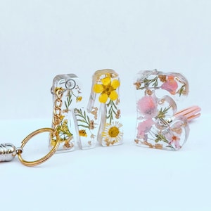 May include: A keychain with a tassel and three resin letters that spell "ME" with pressed flowers and gold leaf inside. The letters are clear and the flowers are yellow, pink, and white.