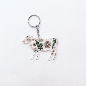 May include: A clear acrylic keychain shaped like a cow with a floral design. The design features green leaves, white flowers, and gold glitter.