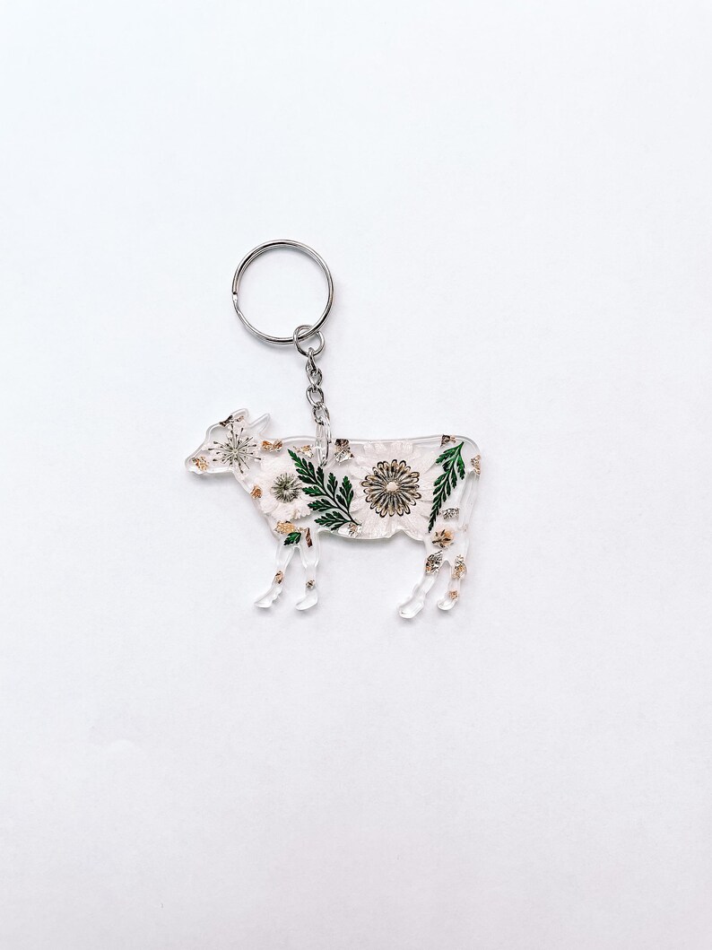 May include: A clear acrylic keychain shaped like a cow with a floral design. The design features green leaves, white flowers, and gold glitter.