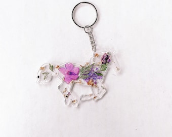 Puede incluir: Un llavero de caballo de resina transparente con flores prensadas de color morado y blanco y purpurina dorada en el interior.