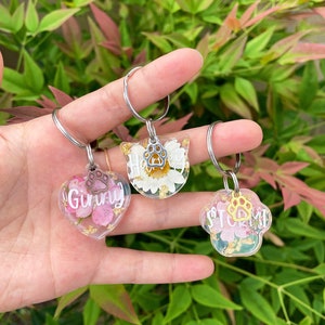 以下が含まれることがあります： 花と金箔を閉じ込めたレジン製のキーホルダー3個。キーホルダーはハート型、肉球型、猫の頭の形をしています。それぞれのキーホルダーには、リングに銀色の肉球チャームが付いています。ハート型のキーホルダーには「Ginny」、肉球型のキーホルダーには「Storm」、猫の頭型のキーホルダーには「Harvey」と書いてあります。