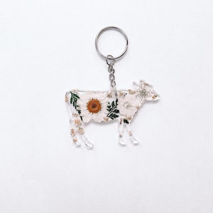 May include: A white cow keychain with pressed flowers and a silver key ring. The cow is made of clear acrylic and has a floral design.