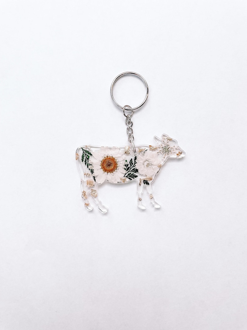 May include: A white cow keychain with pressed flowers and a silver key ring. The cow is made of clear acrylic and has a floral design.