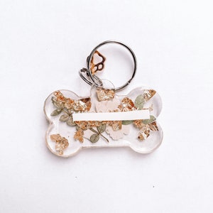 以下が含まれることがあります： 金箔とドライフラワーが入った透明なレジン製の犬の骨の形をしたキーホルダー。キーホルダーにはシルバーのリングと肉球のチャームが付いています。