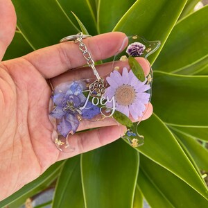 May include: A purple and gold resin dog keychain with a paw print charm and the name "Joey" written on it. The keychain is decorated with pressed flowers.