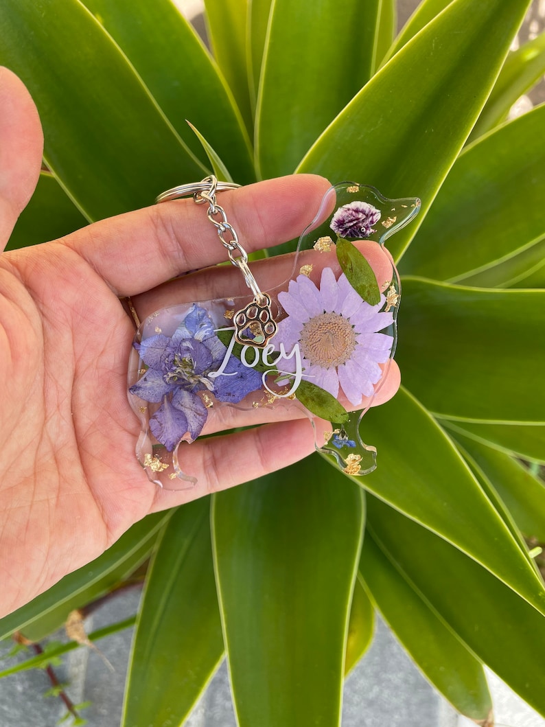 May include: A purple and gold resin dog keychain with a paw print charm and the name "Joey" written on it. The keychain is decorated with pressed flowers.