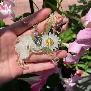 May include: A clear resin keychain shaped like a dog with a paw print charm. The keychain is filled with dried white flowers and gold glitter. The word "Izzy" is written in white on the keychain.