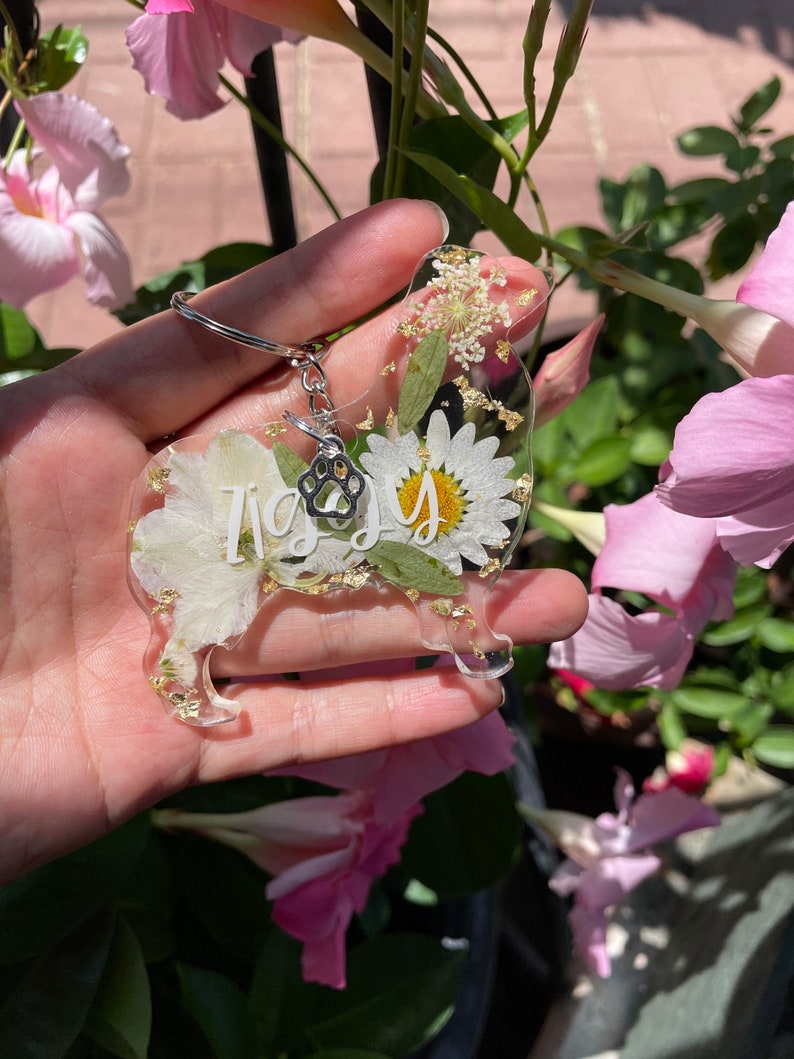 May include: A clear resin keychain shaped like a dog with a paw print charm. The keychain is filled with dried white flowers and gold glitter. The word "Izzy" is written in white on the keychain.