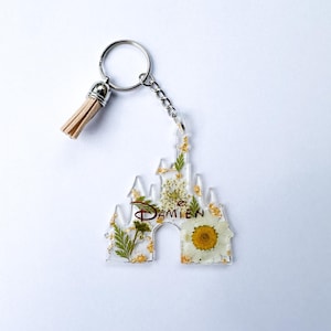 May include: A clear acrylic keychain in the shape of a castle, filled with dried flowers and gold leaf. The name "DAMIEN" is written in a brown script. The keychain has a silver chain, ring, and a tan tassel.