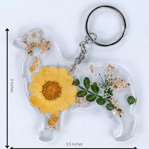 May include: A clear resin keychain shaped like a dog with a yellow daisy, green leaves, and gold flakes embedded inside. The keychain is 3.5 inches wide and 3 inches tall.
