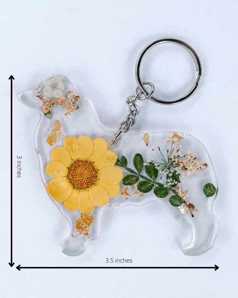 May include: A clear resin keychain shaped like a dog with a yellow daisy, green leaves, and gold flakes embedded inside. The keychain is 3.5 inches wide and 3 inches tall.