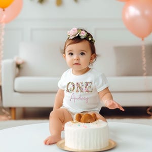 May include: A baby wearing a white t-shirt with the word "ONE" in floral letters and the name "Audrey" in cursive. The baby is sitting in front of a white cake with a teddy bear cake topper. Balloons in shades of pink, orange, and green are in the background.