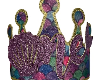 May include: A crown-shaped party hat featuring a colorful scale pattern in shades of purple, blue, and green. The hat is decorated with a large purple glittery seashell and the word one in purple glitter script, all outlined in gold glitter.