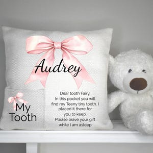 Op de afbeelding: Een witte tandenfee kussen met een roze strik en de naam "Audrey" erop gedrukt. Het kussen heeft een zakje met de woorden "My Tooth" en een kleinere strik. Een witte teddybeer zit naast het kussen.