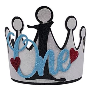 May include: A white crown-shaped party hat featuring a black number one in the center. The word One is written in light blue glittery script across the front, accented by two small red glittery hearts on the left and right sides.