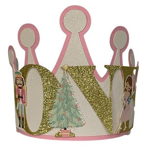 May include: A crown-shaped party hat made of white glittery cardstock with pink trim. The front features the word ONE in large gold glitter letters, a central illustrated Christmas tree, a nutcracker figure, and a girl in a pink dress.