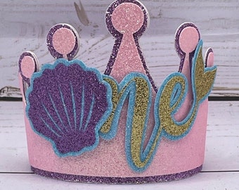 May include: A pink glitter crown with purple and gold accents. The crown features a seashell and the word "one" in a whimsical font, perfect for a first birthday celebration. The crown is decorated with glitter.