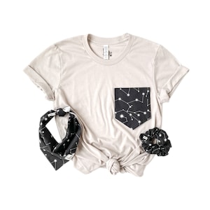 May include: A beige t-shirt with a black pocket featuring a constellation print. The pocket is on the left side of the shirt. The shirt is paired with a black bandana and a black scrunchie, both with the same constellation print.