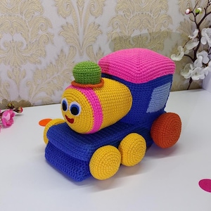 Peut inclure: Un train jouet crocheté aux couleurs vives. La locomotive est jaune avec des détails roses et oranges, le corps est bleu et les roues sont jaunes. Le toit est rose et la cheminée est verte. Le train a un visage souriant.