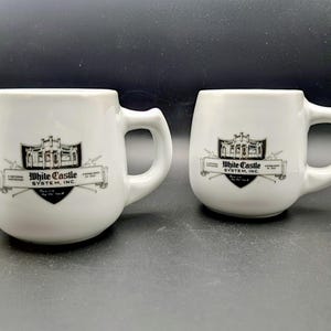 May include: Two white ceramic mugs with rounded bodies and curved handles. Each mug features a black and white graphic of a building and the text "White Castle System, Inc."