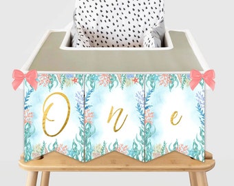 May include: A high chair decorated with a blue and green mermaid-themed banner that says "One" in gold letters. The banner has pink bows on each end.