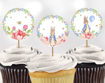 May include: Three cupcakes with white frosting topped with round paper cupcake toppers featuring a watercolor illustration of a bunny rabbit with a blue balloon, flowers, and bees. The bunny is wearing a blue jacket and holding a blue balloon.