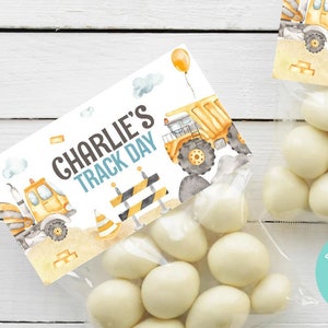 May include: Clear cellophane bags filled with white candies, each topped with a personalized label. The labels feature the text "Charlie's Track Day" with illustrations of construction vehicles, clouds, and traffic cones. A yellow balloon is also visible.