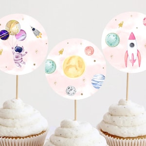 Puede incluir: Tres adornos para cupcakes con diseños espaciales. Uno presenta un astronauta con globos planetarios, otro un sol y planetas, y el último un cohete. Los adornos son redondos con un fondo rosa acuarela y detalles de estrellas doradas.