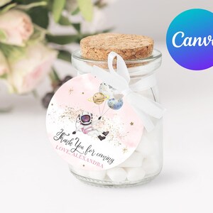 Puede incluir: Un pequeño frasco de vidrio lleno de caramelos blancos y atado con una cinta blanca. Una pegatina redonda rosa y dorada con un astronauta y planetas está pegada al frasco. La pegatina dice "Gracias por venir Con cariño Alexandra".