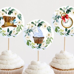 Op de afbeelding: Drie ronde cupcake-toppers met Alice in Wonderland-thema's. De eerste topper heeft een witte en groene bloemenkrans met een bruine en gouden hoed. De tweede topper heeft een witte en groene bloemenkrans met een blauwe en witte cupcake met de woorden "Eat Me". De derde topper heeft een witte en groene bloemenkrans met een gouden zakhorloge met een rood lint.