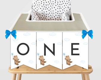 May include: A white banner with blue and white clouds and brown bears holding blue balloons with the number "1" on them. The banner says "ONE" in black letters. The banner is attached to a high chair with blue bows on each end.