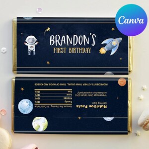 Puede incluir: Un envoltorio de chocolate azul y dorado con temática espacial. Presenta un astronauta, un cohete, estrellas y planetas. El texto en el envoltorio dice "Brandon's First Birthday".